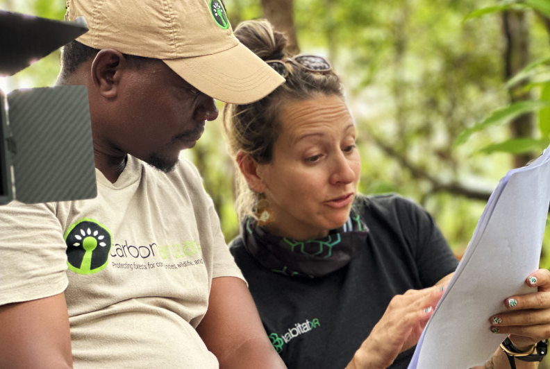 Two people outdoors, one wearing a beige cap and shirt with a green tree logo, discussing and pointing at a document.