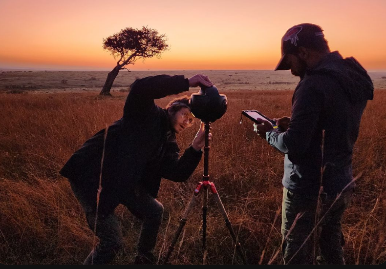 Two men working with a 360 camera mounted on a tripod in a grassy field at sunset with a lone tree in the background.